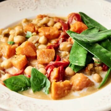 A white plate contains a stew with chunks of sweet potato, chickpeas, red bell pepper, and spinach in a creamy sauce, topped with snow peas. The plate has a decorative, lacy edge.