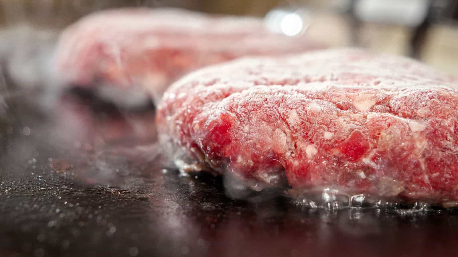 Two frozen raw beef burger patties are sizzling on a hot grill, with steam rising around them as they begin to cook.