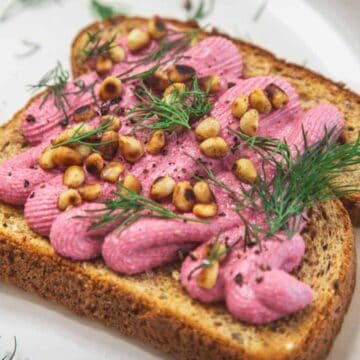 Two slices of whole grain toast topped with piped pink spread, toasted pine nuts, and fresh dill, served on a white plate.