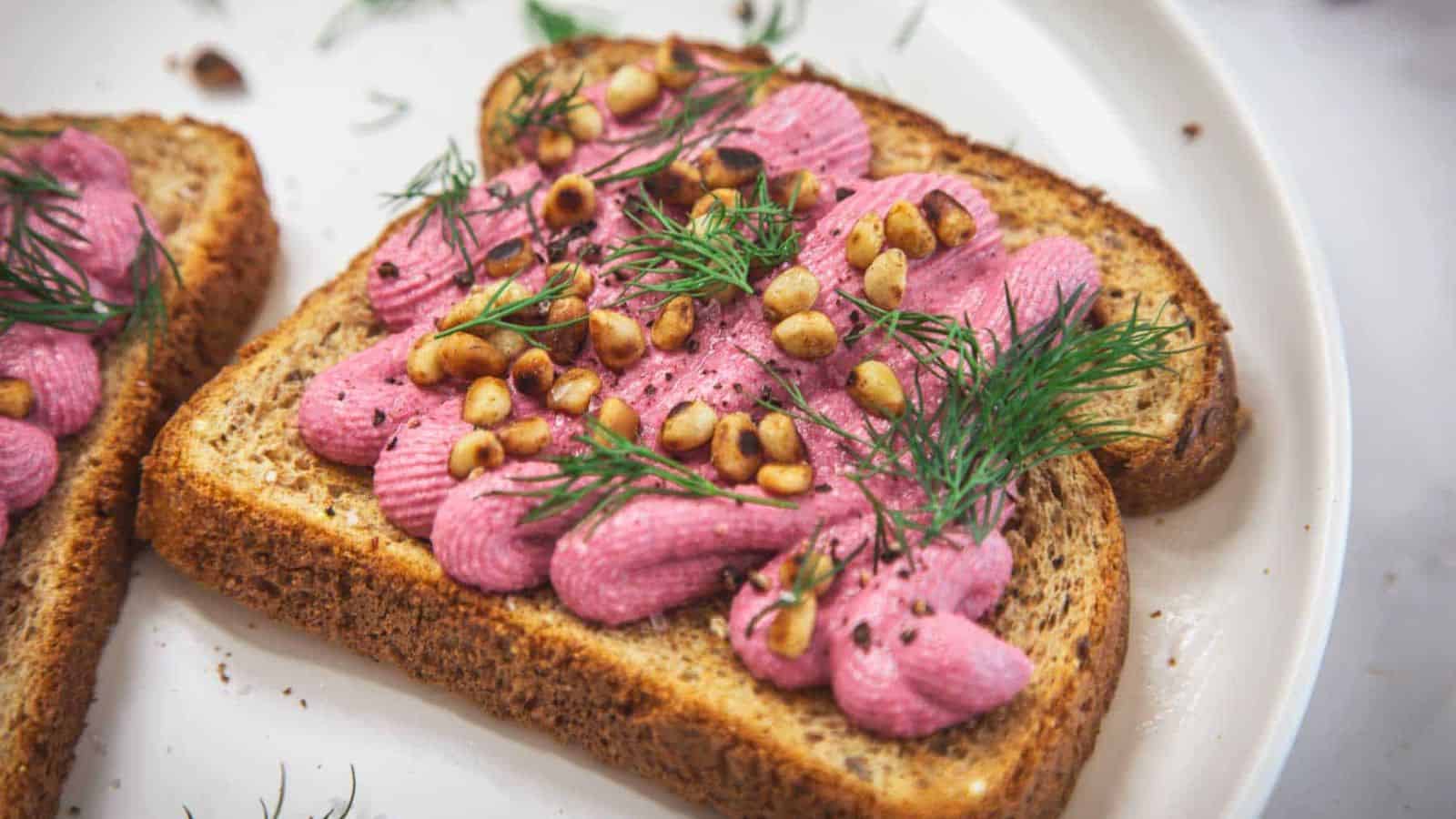 Two slices of whole grain toast topped with piped pink spread, toasted pine nuts, and fresh dill, served on a white plate.