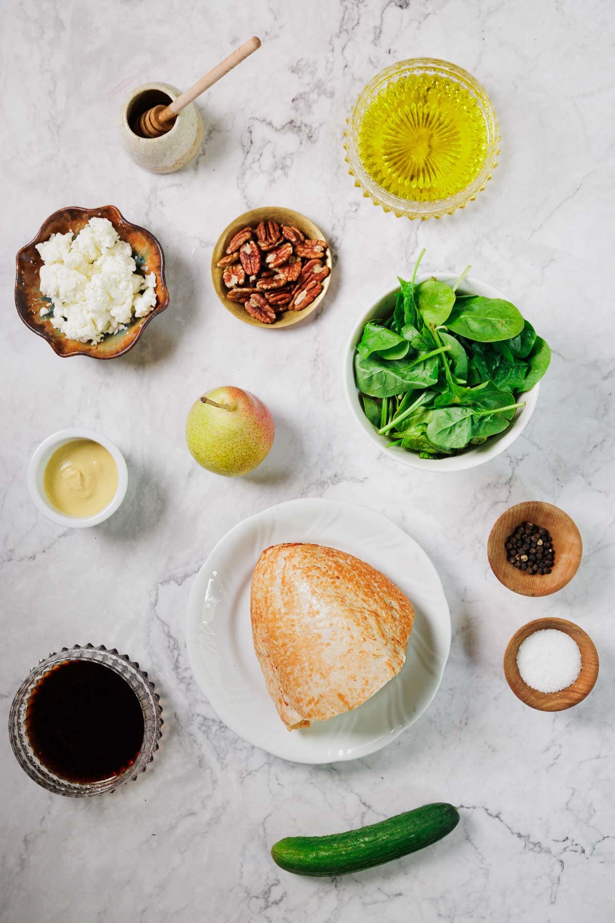 A variety of ingredients arranged on a marble surface, including a bowl of spinach, a block of cheese, a green apple, pecans, cucumber, honey, oil, vinegar, mustard, salt, pepper, and crumbled cheese.