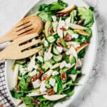 A white oval dish contains a salad with spinach leaves, sliced pears, cucumber, chunks of white cheese, pecans, and pieces of chicken. Two wooden salad servers rest on the edge of the dish.
