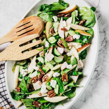 A white oval dish contains a salad with spinach leaves, sliced pears, cucumber, chunks of white cheese, pecans, and pieces of chicken. Two wooden salad servers rest on the edge of the dish.