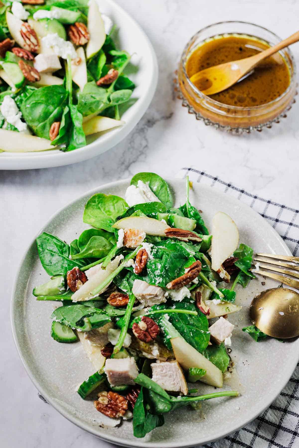 A plate of spinach salad with sliced pears, chicken, pecans, and cheese crumbles. A bowl of the same salad is in the background. A small jar of vinaigrette and a gold fork and spoon are beside the plate.