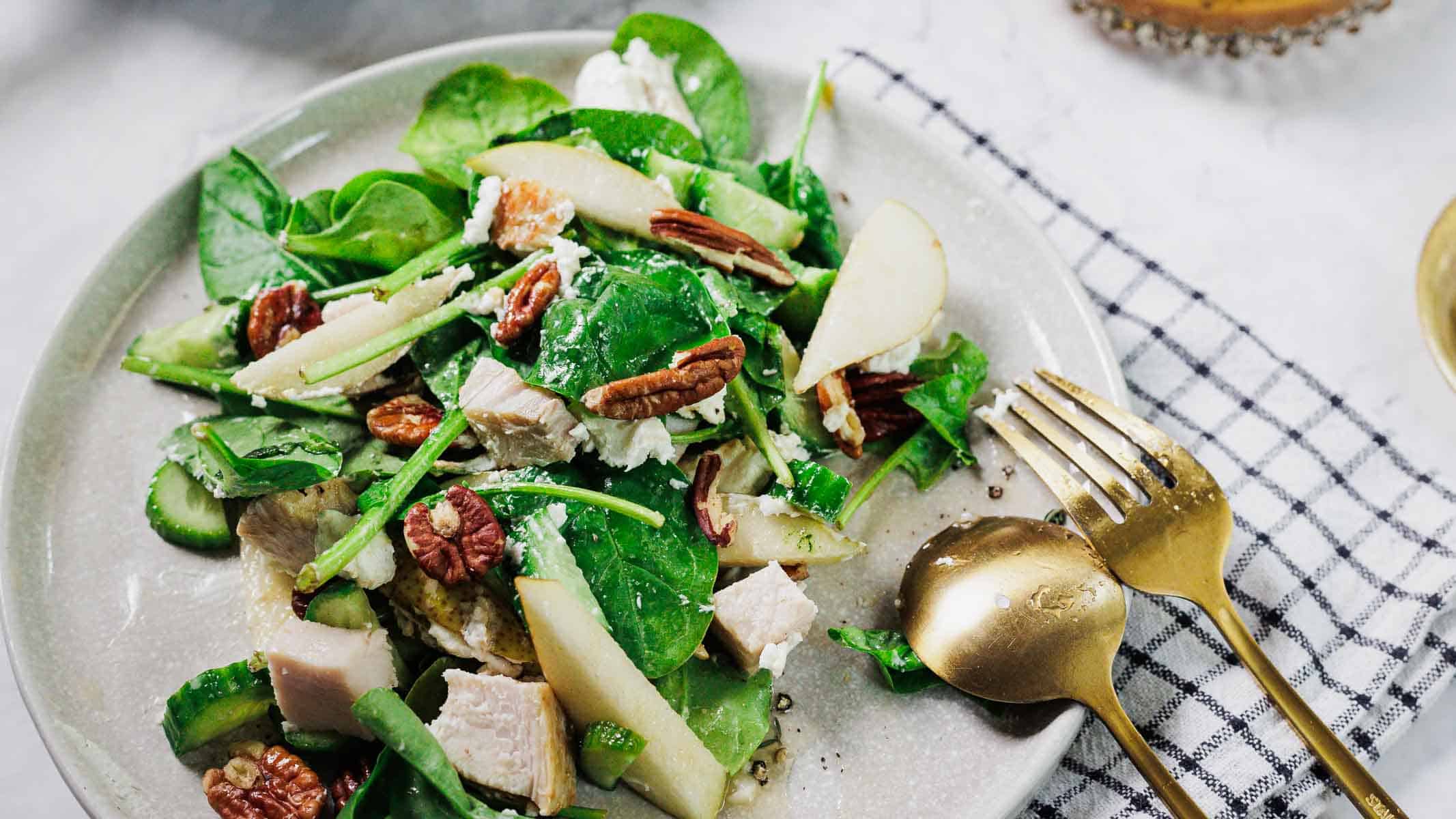 A plate of salad with spinach, pecans, pear slices, cucumber, and chunks of white meat, served on a white plate with a gold fork and spoon on a checkered napkin.