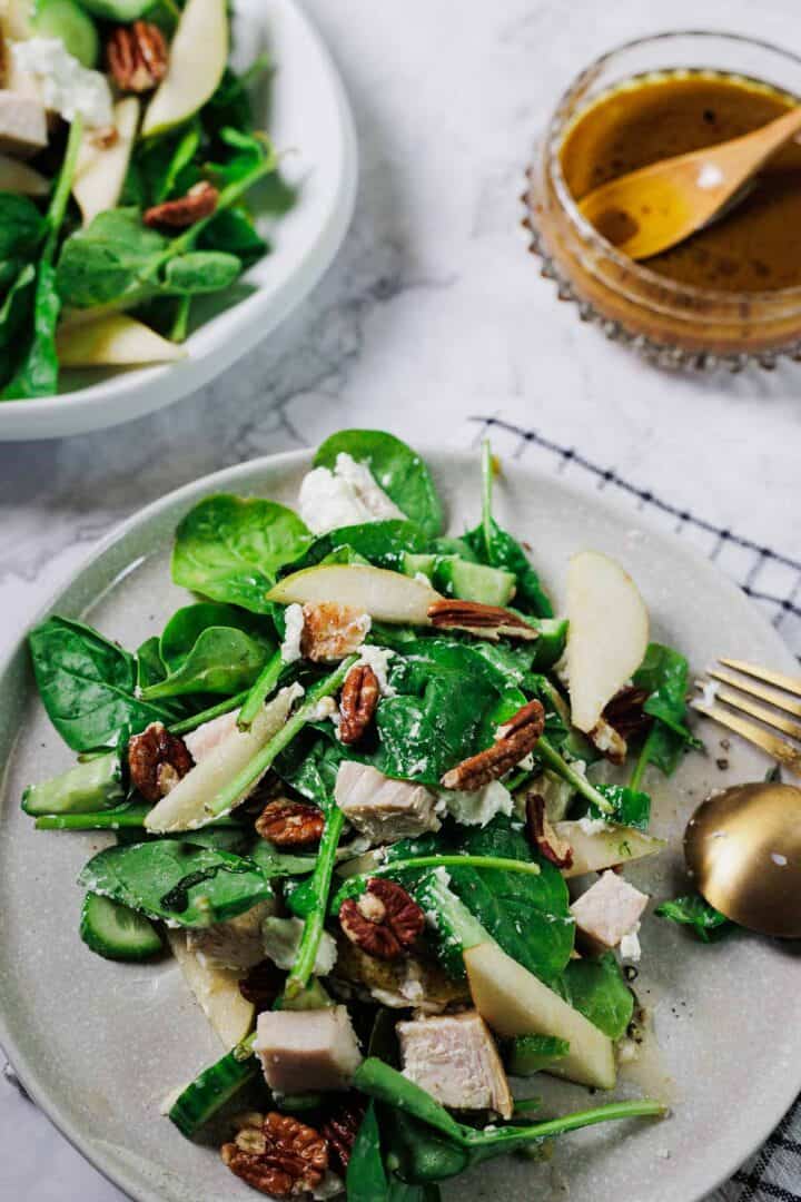 A plate of salad with spinach, sliced pears, chicken, pecans, cucumber, and feta cheese sits next to a bowl of vinaigrette dressing on a marble surface with a gold fork beside the plate.