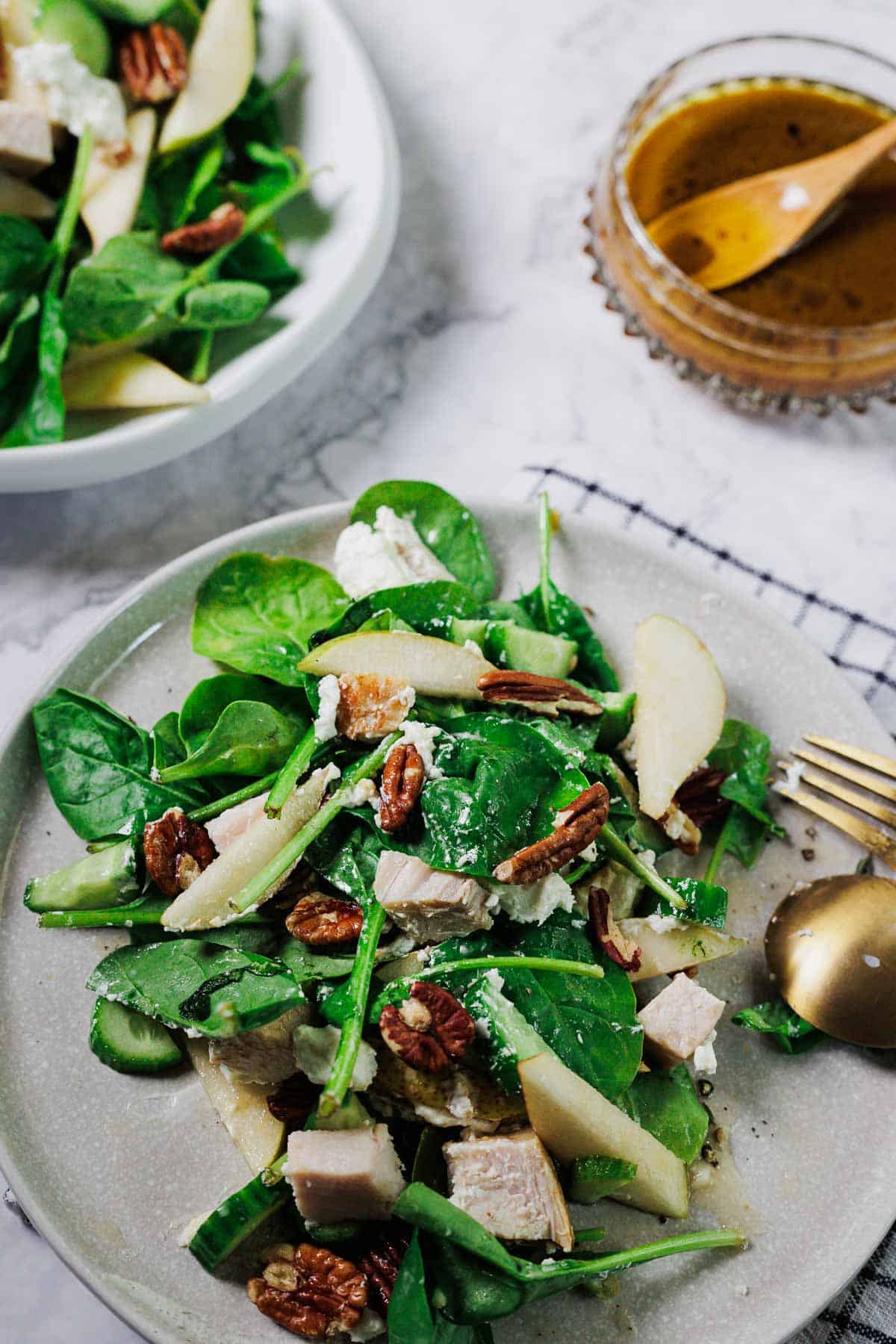 A plate of salad with spinach, sliced pears, chicken, pecans, cucumber, and feta cheese sits next to a bowl of vinaigrette dressing on a marble surface with a gold fork beside the plate.