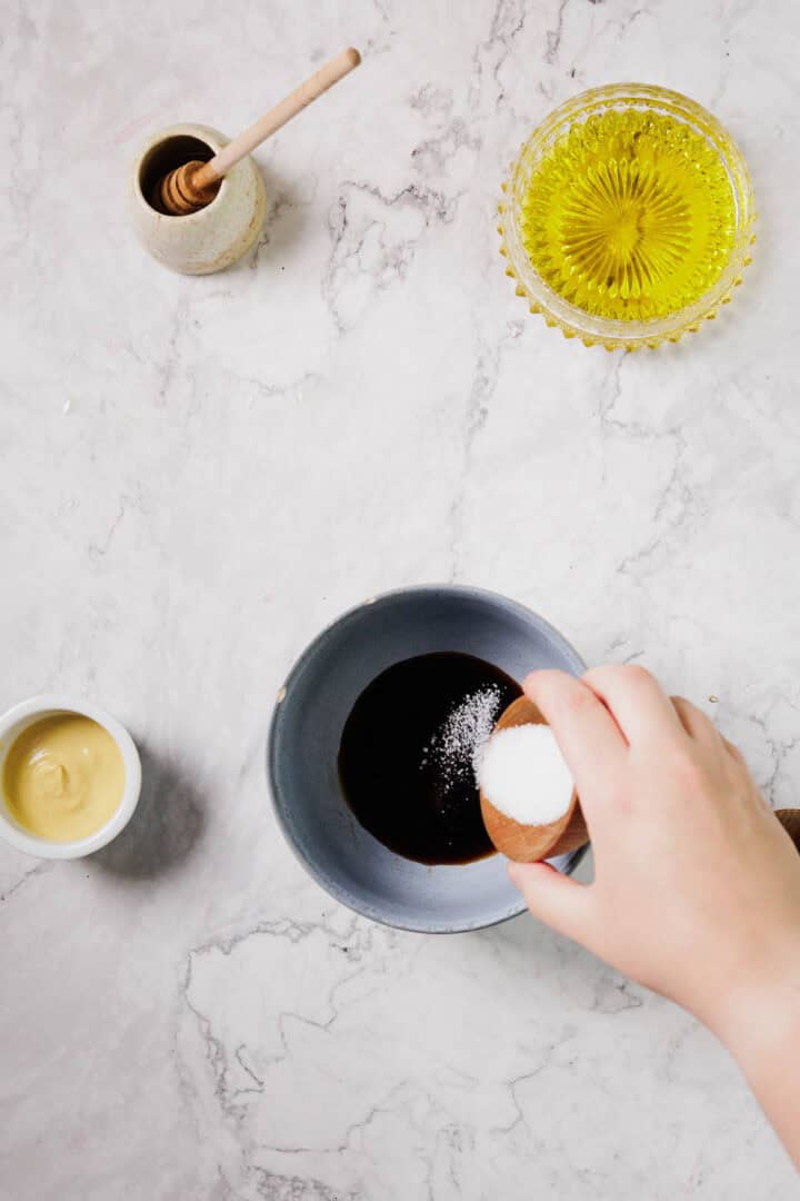 A hand pours salt into a bowl of dark liquid, with small bowls of mustard, honey, and olive oil arranged on a light marble countertop.