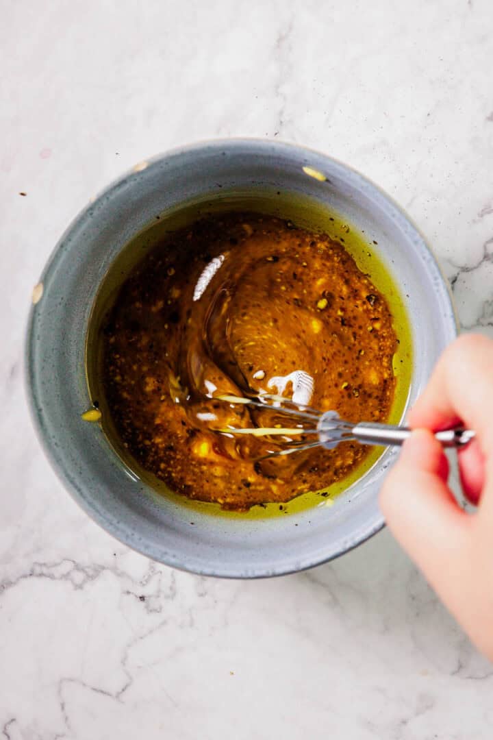 A hand using a small whisk to mix a golden-brown liquid with visible spices in a gray bowl on a marble countertop.