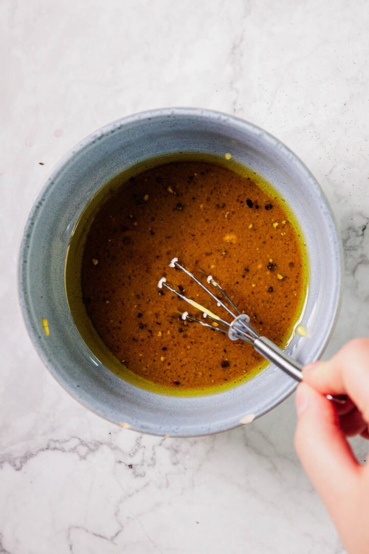 A hand holds a whisk stirring a yellow-brown liquid with visible pepper flakes in a gray bowl on a white marble surface.