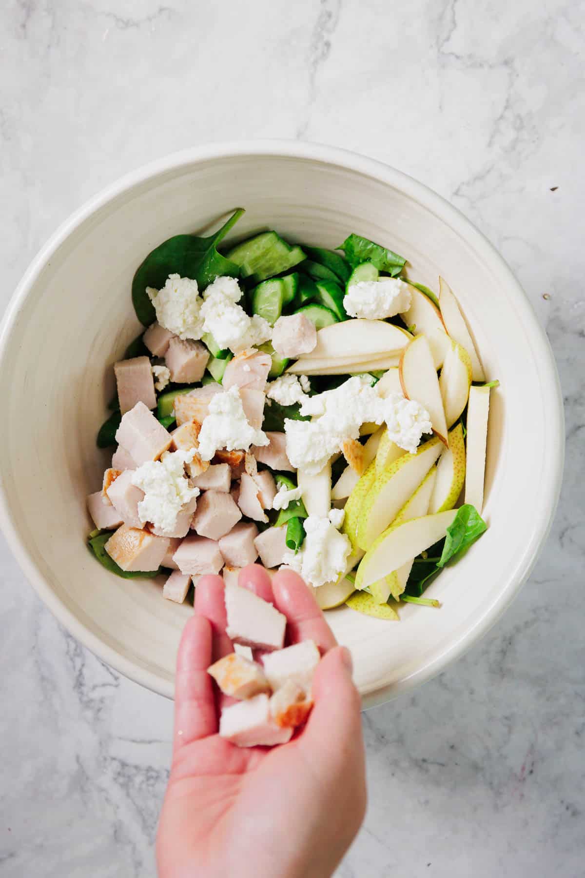 A hand holds cubed cooked chicken above a bowl containing spinach, sliced cucumber, sliced pear, and dollops of white cheese on a white marble surface.