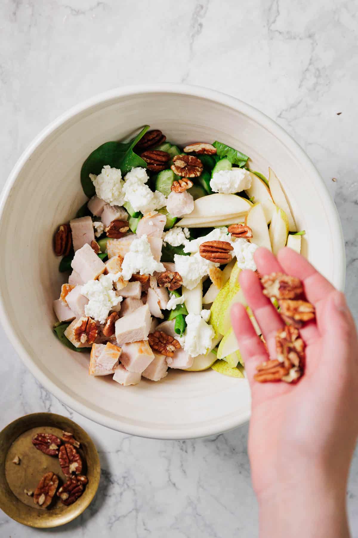 A hand adds pecans to a salad in a white bowl. The salad contains spinach, sliced pears, diced chicken, goat cheese, and more pecans on a marble surface. A small brass bowl with pecans sits nearby.