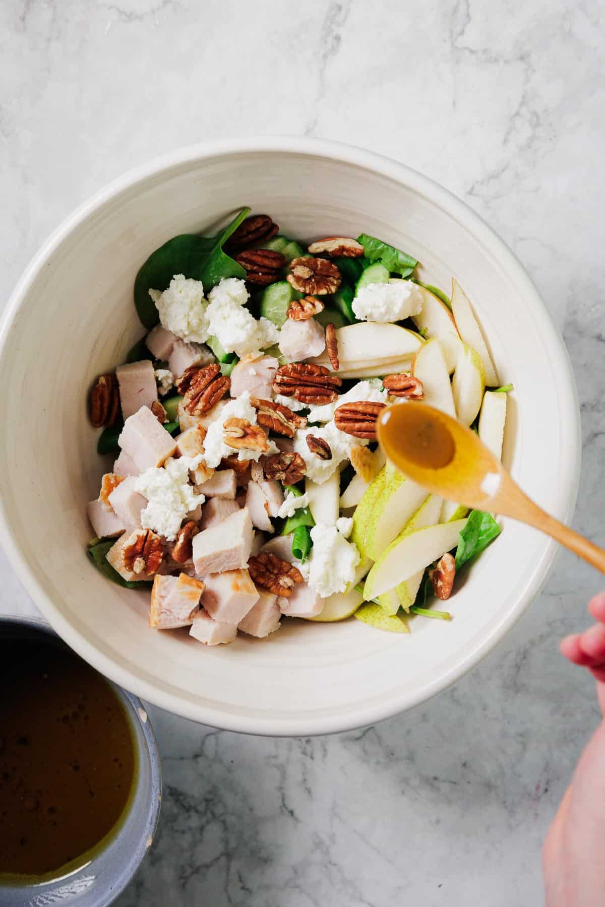 A hand holds a spoon of dressing above a bowl containing spinach, sliced pears, cubed chicken, pecans, and crumbled goat cheese. A small bowl of extra dressing sits nearby on a marble surface.