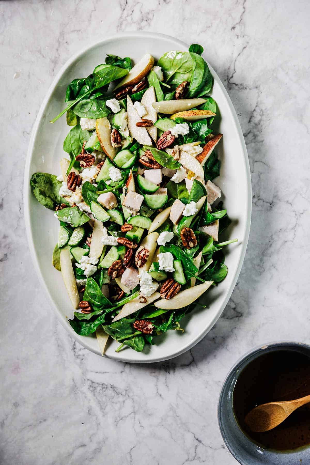 An oval white plate holds a spinach salad with sliced pears, pecans, crumbled cheese, and chunks of chicken. A small bowl with a wooden spoon sits nearby on a marble surface.