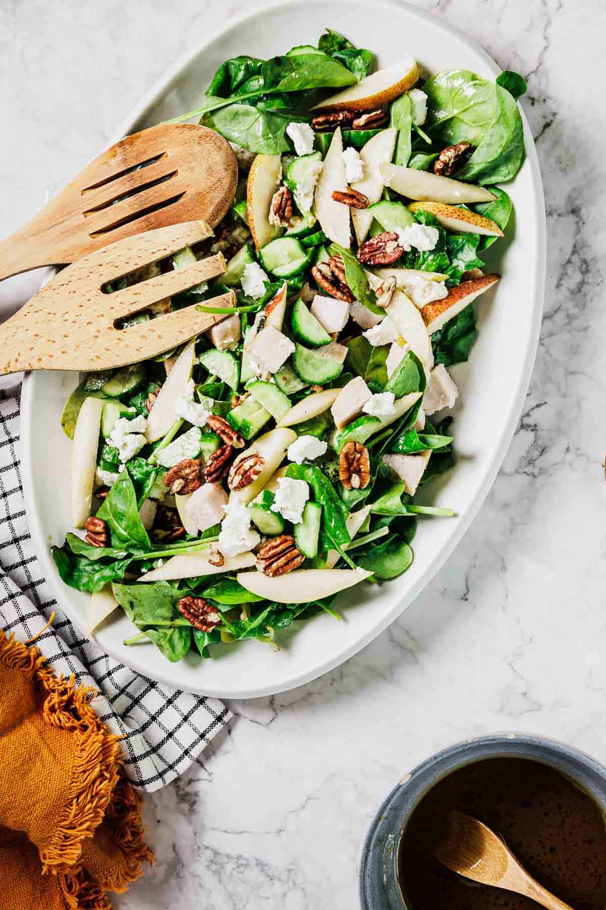 A white oval plate holds a spinach salad with sliced pears, pecans, cucumber, diced chicken, and crumbled cheese. Wooden salad tongs rest on top, and a cup of dressing with a spoon sits nearby on a marble surface.