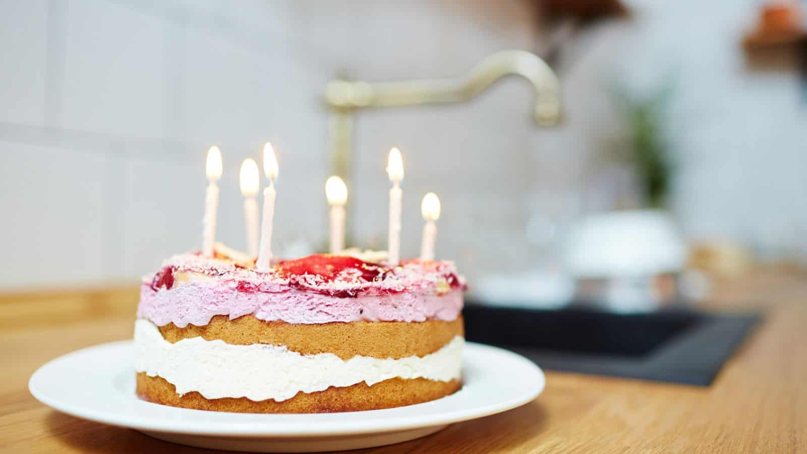 A round cake with white and pink frosting, topped with five lit candles, sits on a white plate on a wooden counter. A kitchen sink and tiled wall are visible in the blurred background.
