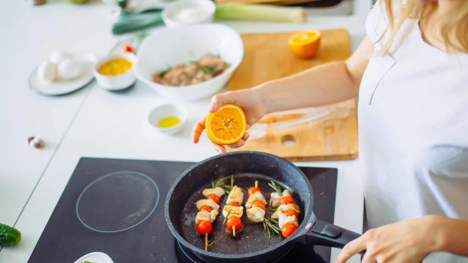 A person in a white shirt cooks kebabs with cherry tomatoes and herbs in a frying pan while holding half an orange over the pan. Various cooking ingredients are on the counter in the background.