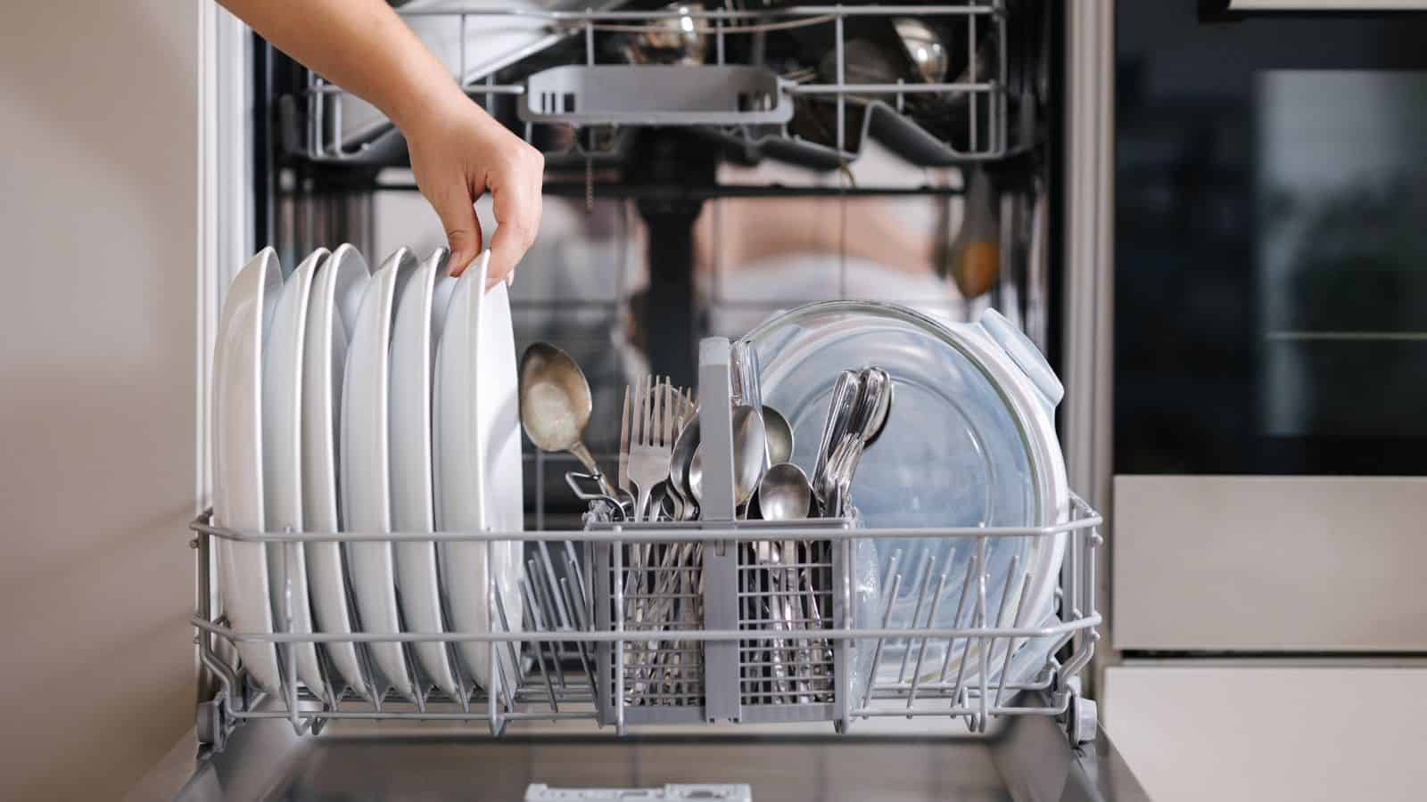 A person’s hand places a white plate into the bottom rack of an open dishwasher loaded with plates, bowls, and various utensils. The dishwasher door is open, and the kitchen setting is visible.