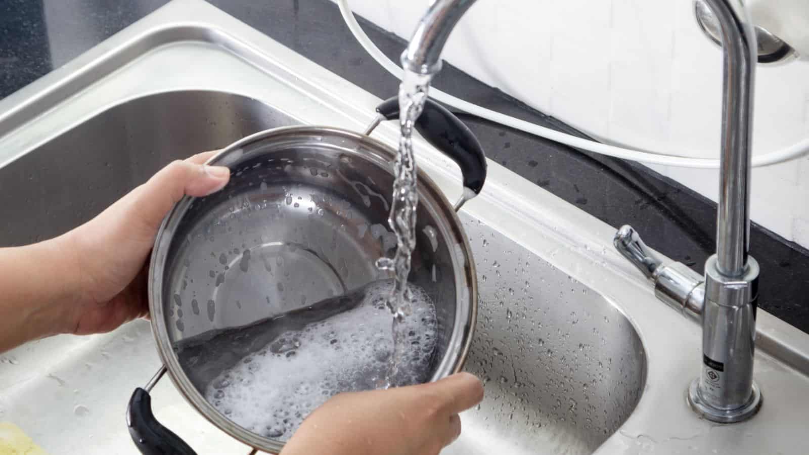A person is washing a soapy stainless steel pot under running water at a kitchen sink. The pot is being held with both hands, and water is flowing from the faucet.
