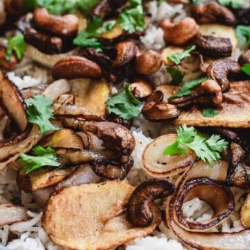 A close-up of a bowl filled with white rice, topped with caramelized onions, fried potato slices, toasted cashews, mushrooms, and fresh cilantro leaves.