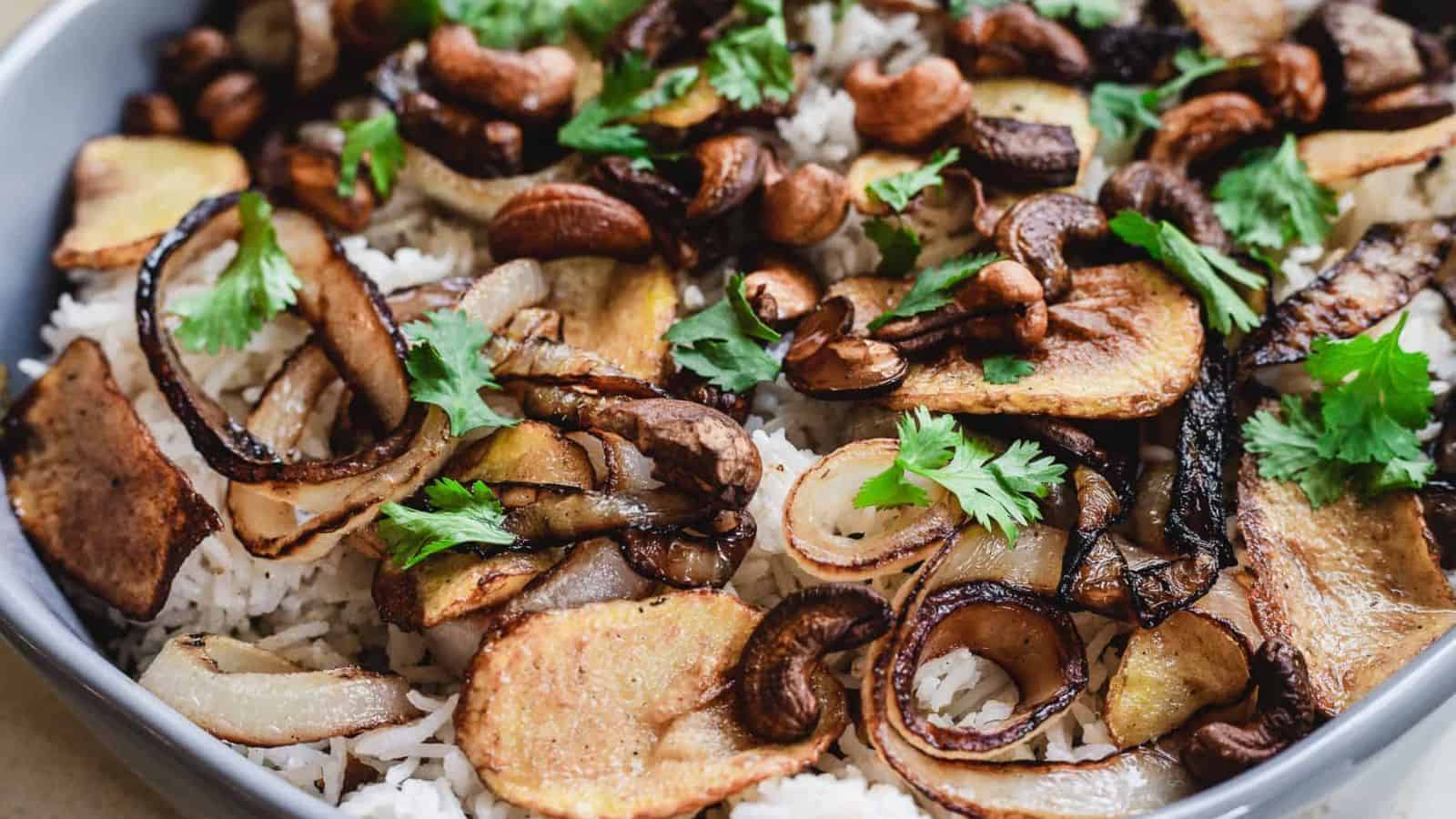 A close-up of a bowl filled with white rice, topped with caramelized onions, fried potato slices, toasted cashews, mushrooms, and fresh cilantro leaves.