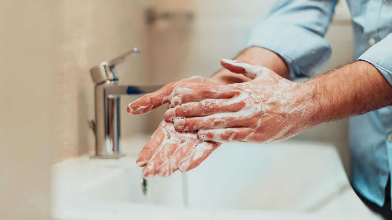 A person is washing their hands at a sink, rubbing their hands together with soap to create lather. The faucet is on, and the individual is wearing a light blue long-sleeve shirt.