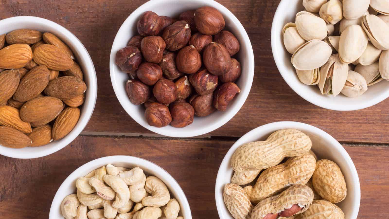 Five small white bowls filled with different types of nuts, including almonds, hazelnuts, pistachios, cashews, and peanuts, arranged on a wooden surface.