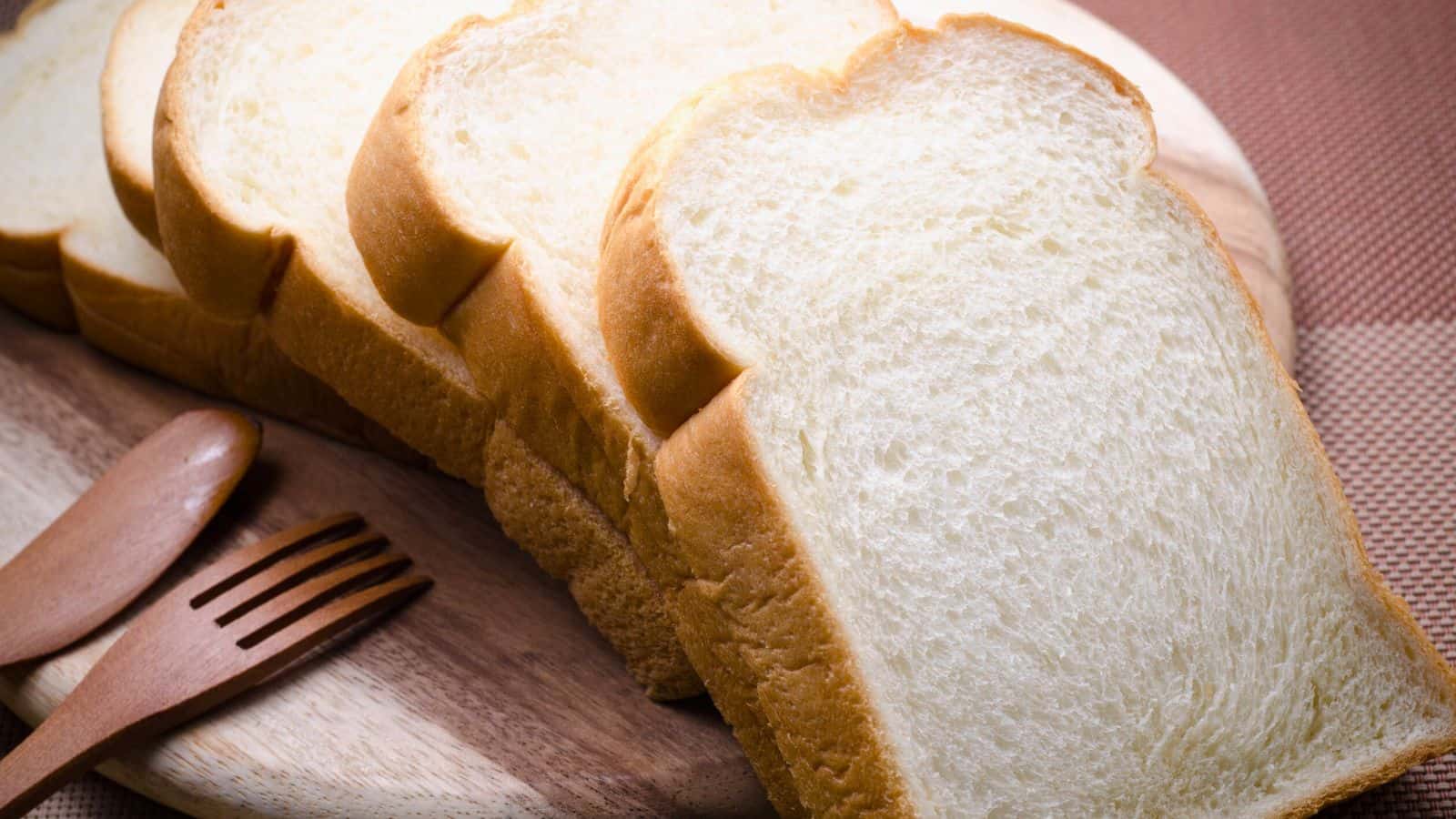 Several slices of white bread are arranged on a wooden cutting board next to a wooden fork and spoon, all set on a brown textured surface.