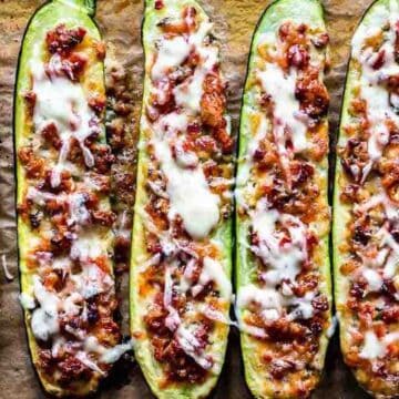 Six zucchini halves are arranged in a row on parchment paper, filled with a browned meat and tomato mixture, and topped with melted white cheese.