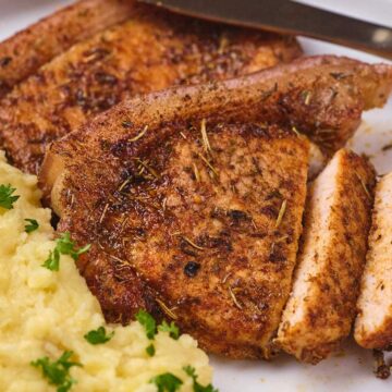 A plate with seasoned and cooked pork chops, some sliced, next to a serving of mashed potatoes garnished with chopped parsley. A knife rests at the top of the plate.