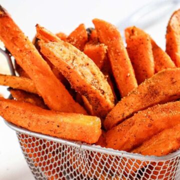 A metal basket filled with seasoned sweet potato fries sits on a white surface. A bowl of red dipping sauce is partially visible in the background. The fries are golden-orange and coated with spices.
