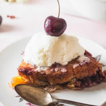 A slice of cherry cobbler topped with vanilla ice cream and a cherry, served on a white plate with a spoon—perfect for desserts to impress your in-laws. Scattered cherries complete this tempting treat, inspired by classic recipes.