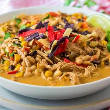 A white bowl filled with creamy soup containing shredded chicken, black beans, corn, and white beans, topped with red, blue, and yellow tortilla strips, sliced jalapeños, and a lime wedge.