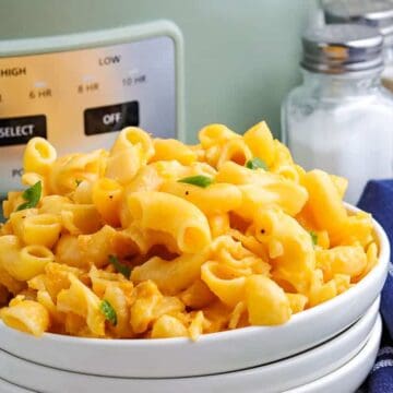 A white bowl filled with macaroni and cheese sits on a table next to a blue striped napkin, salt and pepper shakers, a sprig of parsley, and a slow cooker in the background.