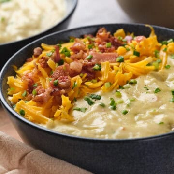 A bowl of creamy potato soup topped with shredded cheddar cheese, bacon pieces, and chopped chives sits on a wooden surface next to a beige cloth napkin. Another bowl of soup is visible in the background.