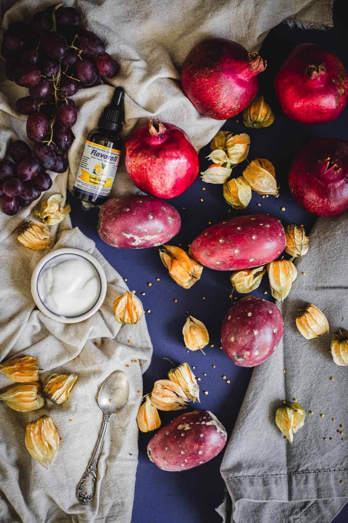 A flat lay of pomegranates, a prickly pear smoothie bowl, cape gooseberries, a bunch of grapes, a spoon, and a dropper bottle arranged on fabric napkins over a dark surface.