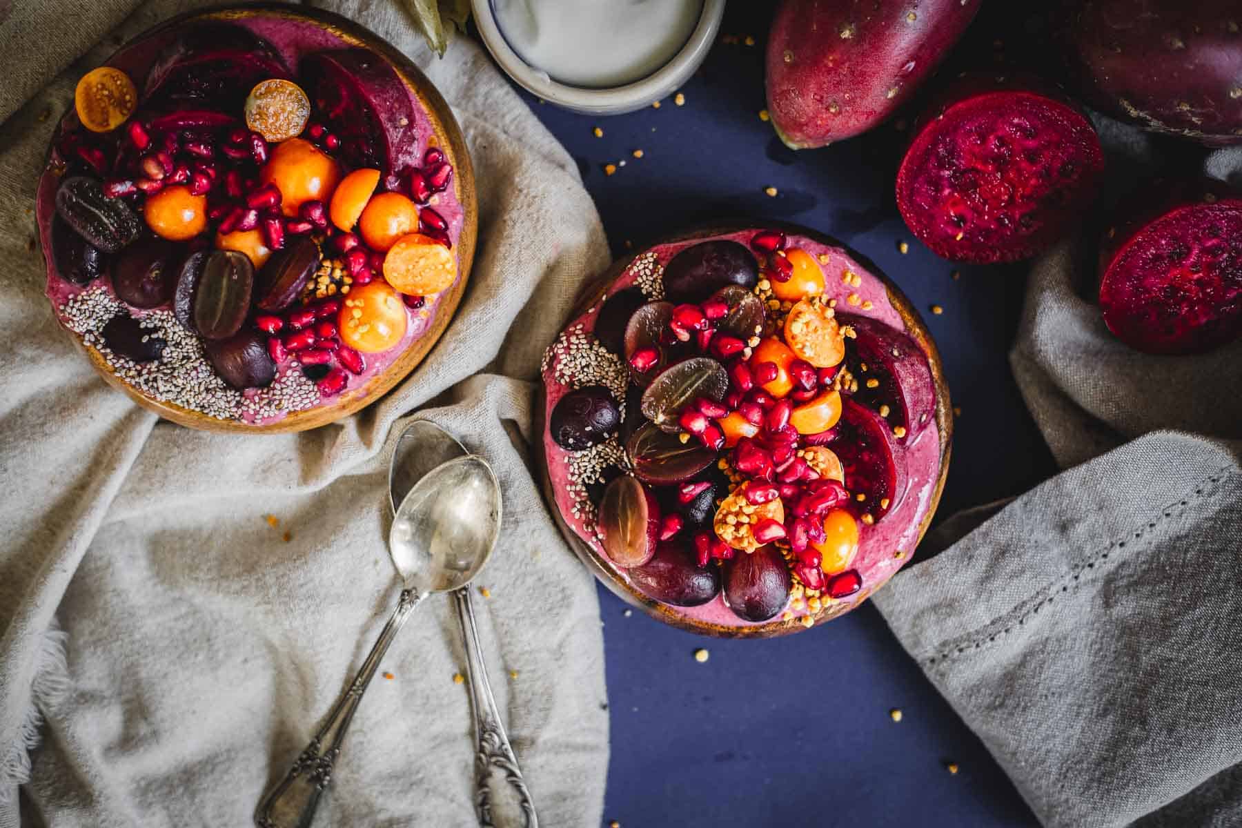 Two Prickly Pear Smoothie Bowls topped with sliced grapes, physalis, pomegranate seeds, chia seeds, and other fresh fruits rest on a dark blue surface beside two spoons, napkins, and vibrant halved dragon fruit.