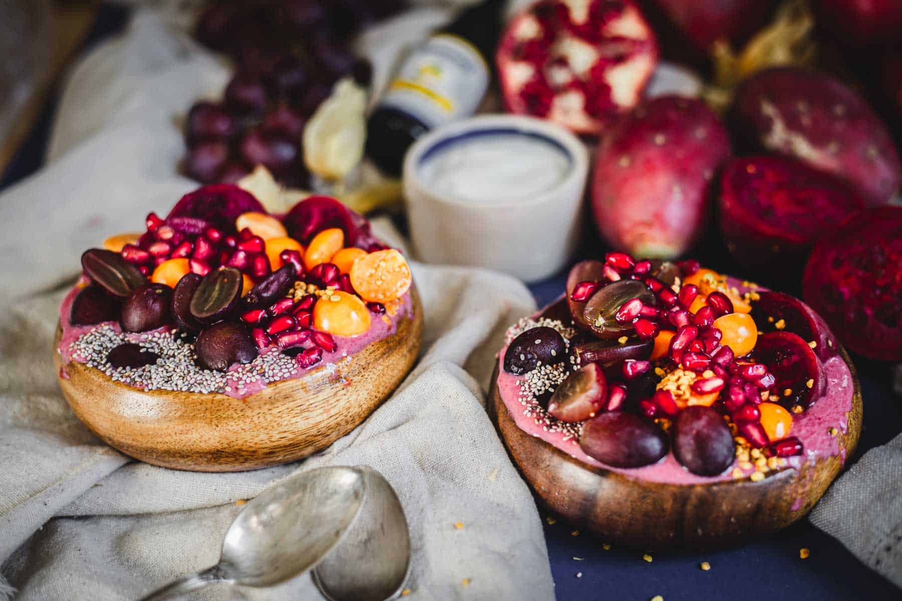 Two wooden bowls filled with a prickly pear smoothie bowl, topped with grapes, pomegranate seeds, cape gooseberries, beet slices, and chia seeds. A small bowl of yogurt, pomegranate halves, and grapes are visible in the background.