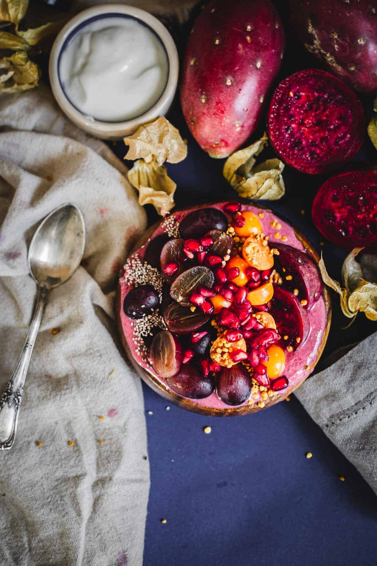 A Prickly Pear Smoothie Bowl topped with grapes, pomegranate seeds, cape gooseberries, and seeds sits on a dark surface next to a spoon, a cloth, a bowl of yogurt, and whole prickly pears.