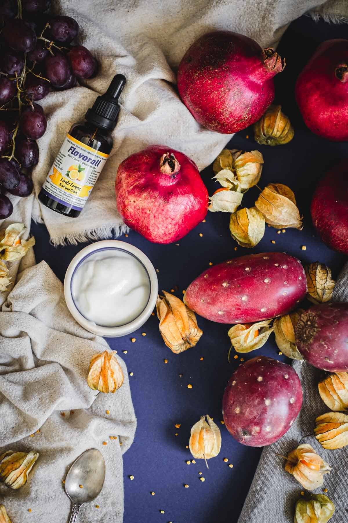 A flatlay of pomegranates, purple grapes, prickly pears, golden berries, a bottle of liquid supplement, and a small bowl of yogurt on a navy surface with a beige cloth and spoon&mdash;perfect inspiration for your next Prickly Pear Smoothie Bowl.