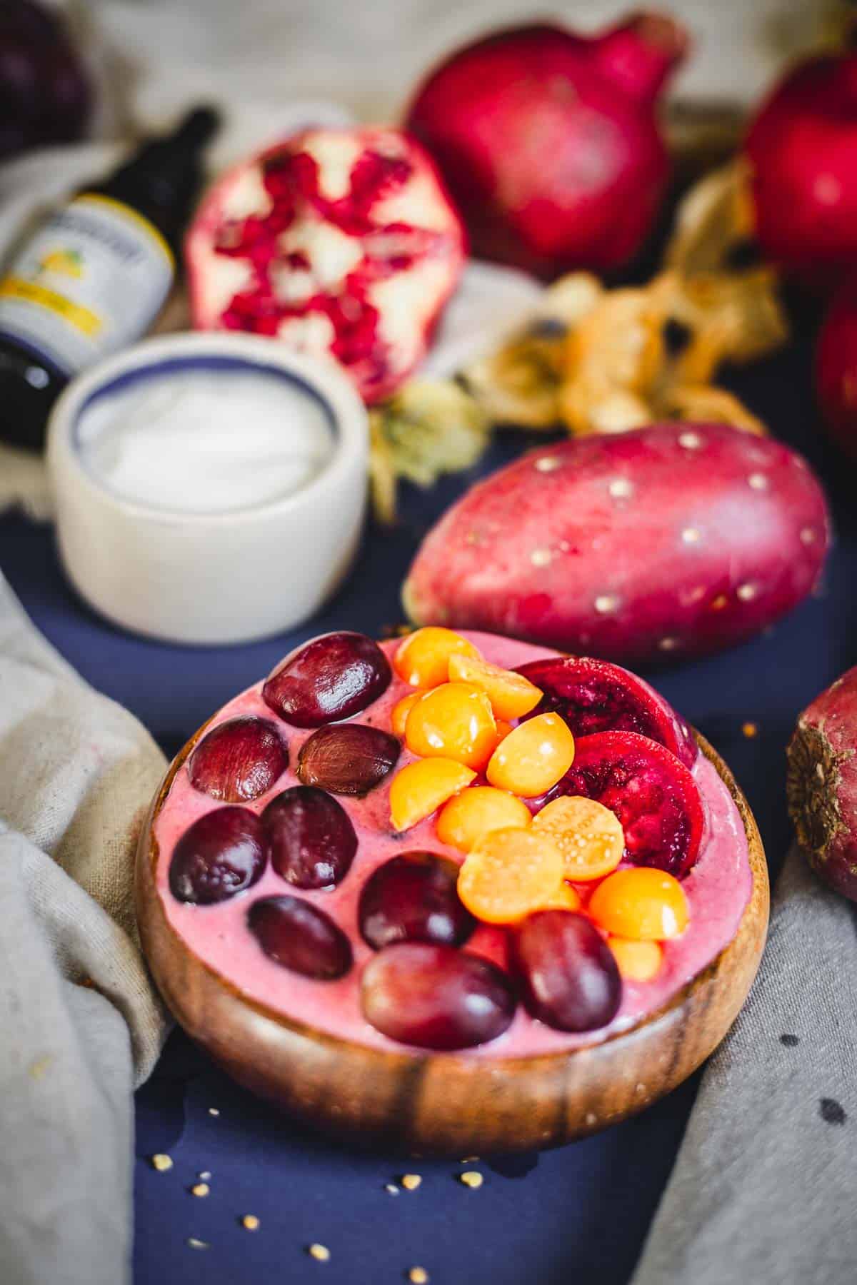 A wooden bowl with a pink Prickly Pear Smoothie Bowl topped with grapes, golden berries, and sliced plums sits on a cloth next to prickly pears, a halved pomegranate, a jar of yogurt, and a small bottle.