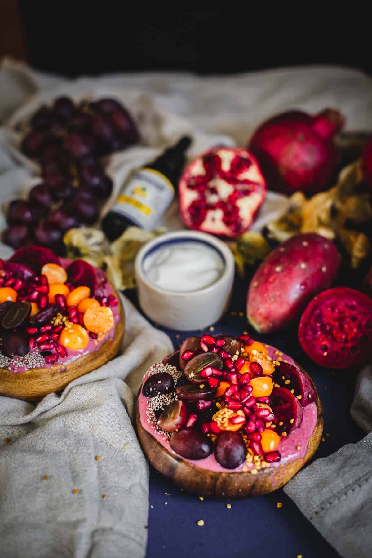 A Prickly Pear Smoothie Bowl topped with pomegranate seeds, grapes, and orange berries sits on a cloth beside creamy yogurt, a halved pomegranate, grapes, and a bottle, with assorted fruits scattered invitingly around.
