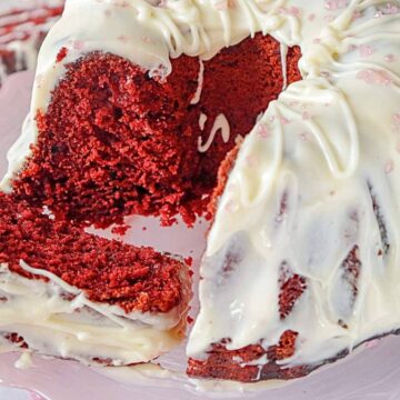 A red velvet bundt cake with white icing is displayed on a pink cake stand. A large slice has been cut and placed in front of the cake, showing its moist, red interior. Pink sprinkles are scattered on the icing.