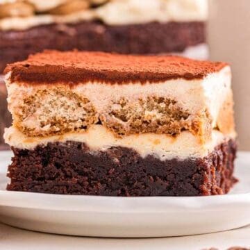 A slice of dessert on a white plate, featuring a layer of brownie on the bottom, creamy filling in the middle, and coffee-soaked ladyfingers dusted with cocoa powder on top.