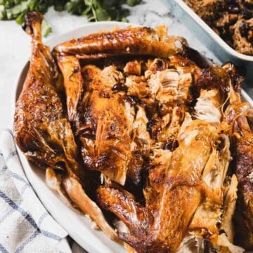 A platter with a roasted turkey wrapped in cheesecloth, carved to reveal golden-brown skin and juicy meat, placed next to a checkered cloth and fresh green herbs.