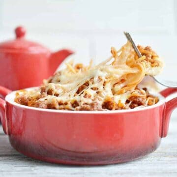 A red ceramic dish filled with baked spaghetti topped with melted cheese and ground meat. A fork is lifting a portion of the pasta. A red teapot and a patterned cloth are in the background.