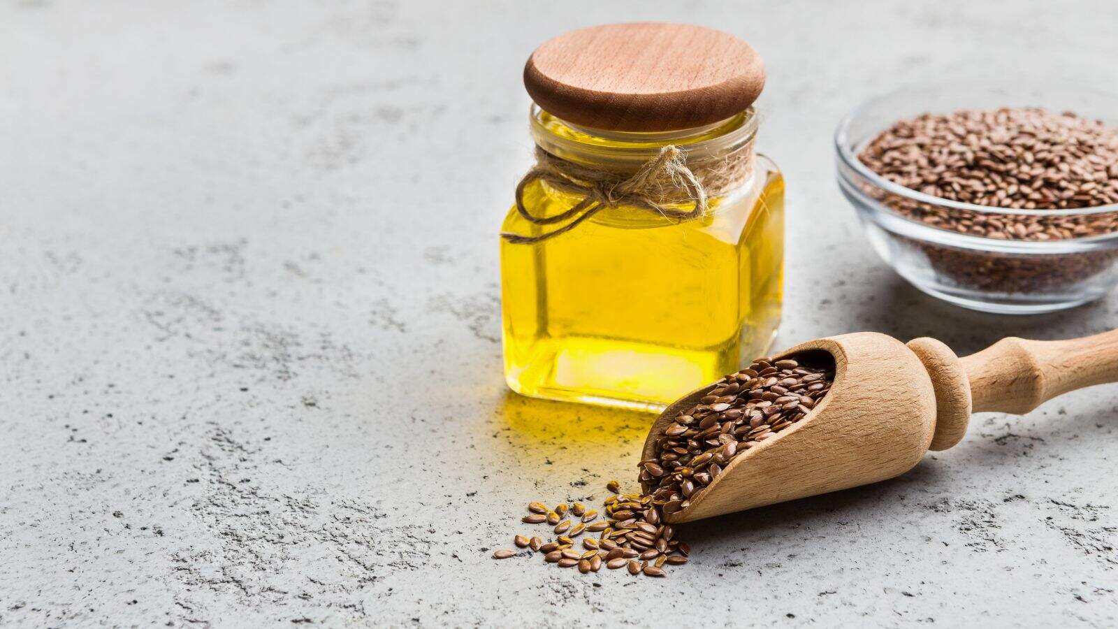 A small glass jar filled with yellow oil and sealed with a wooden lid sits next to a clear bowl of brown seeds and a wooden scoop spilling seeds onto a light gray surface.