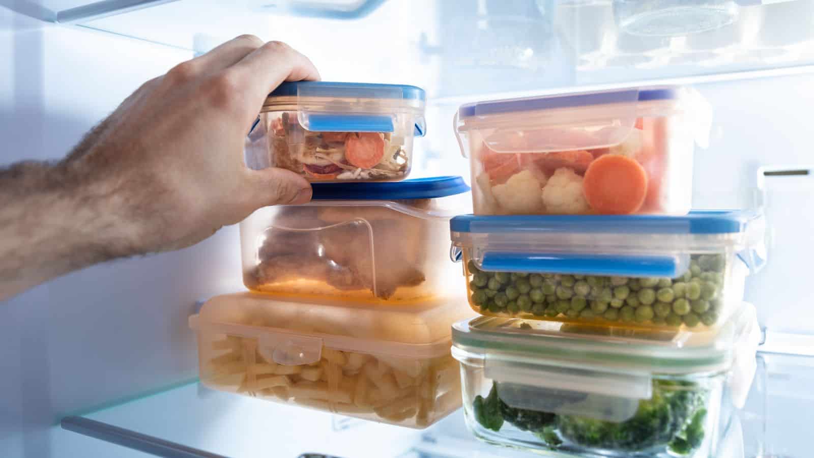 A hand reaching into a refrigerator to place or retrieve a plastic container with food. Several other containers with various foods, including vegetables and what appears to be pasta, are stacked inside the fridge.