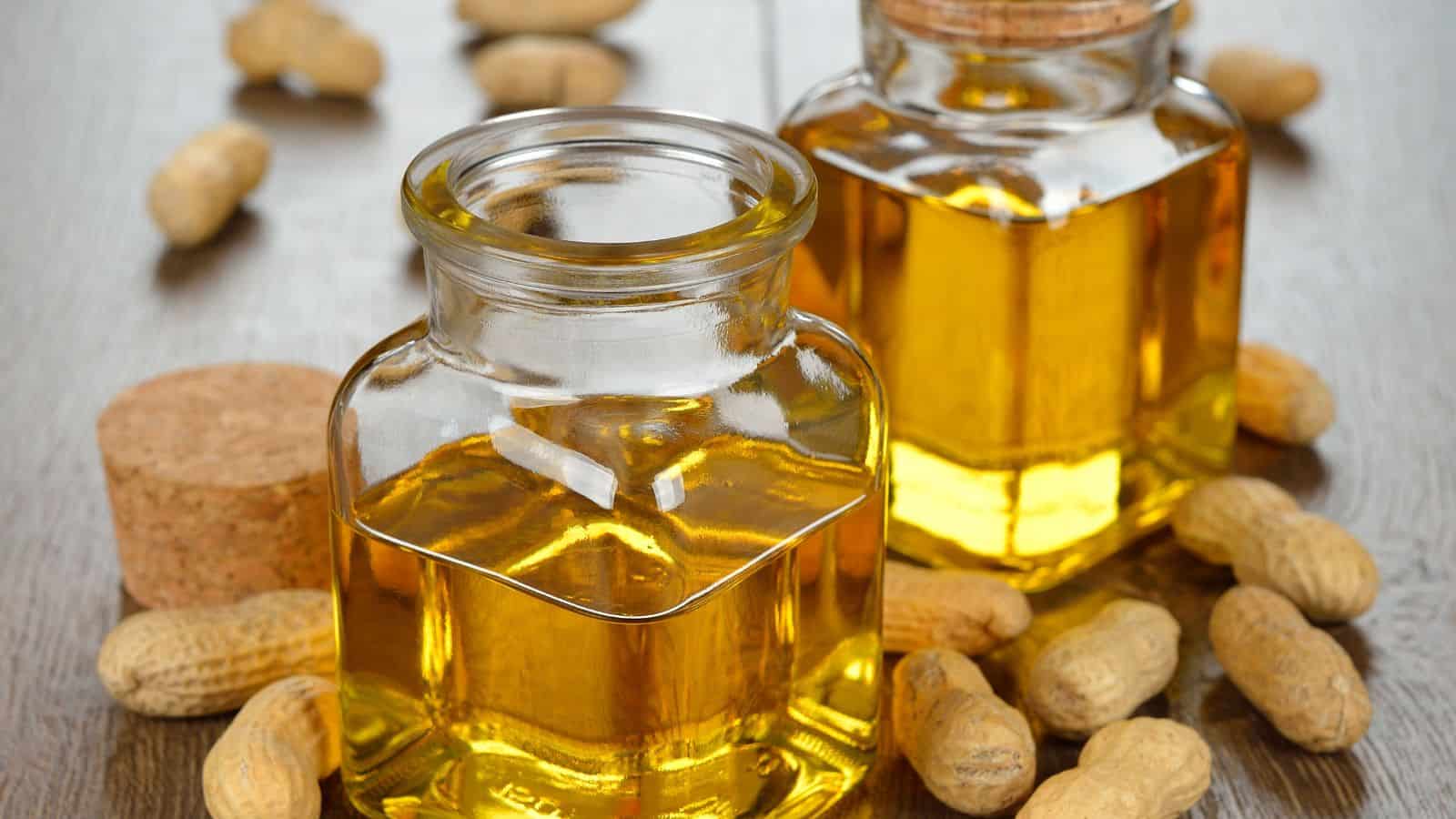 Two glass jars filled with yellow oil are placed on a wooden surface, surrounded by unshelled peanuts. One jar has a cork stopper beside it, and the other jar is closed.