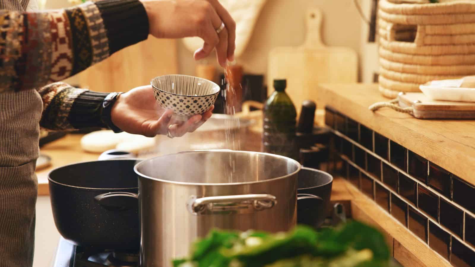 A person sprinkles seasoning into a large metal pot on a stove, holding a small patterned bowl in one hand. The kitchen counter and utensils are visible in the background.
